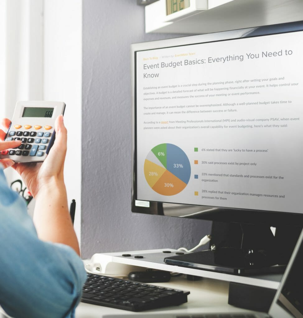 A woman calculating a budget on her calculator while a blog post about budgeting is shown on her desktop monitor.