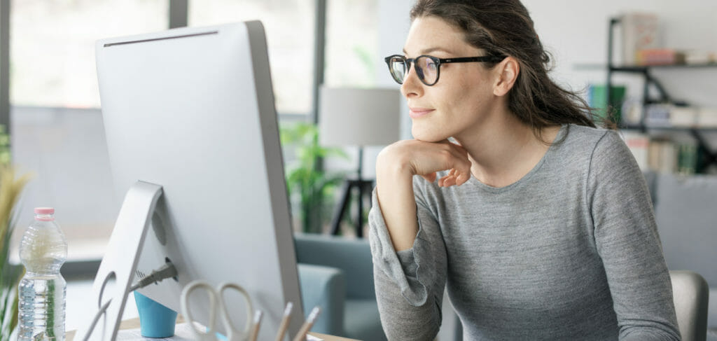 A woman with glasses looking at a computer screen while grinning