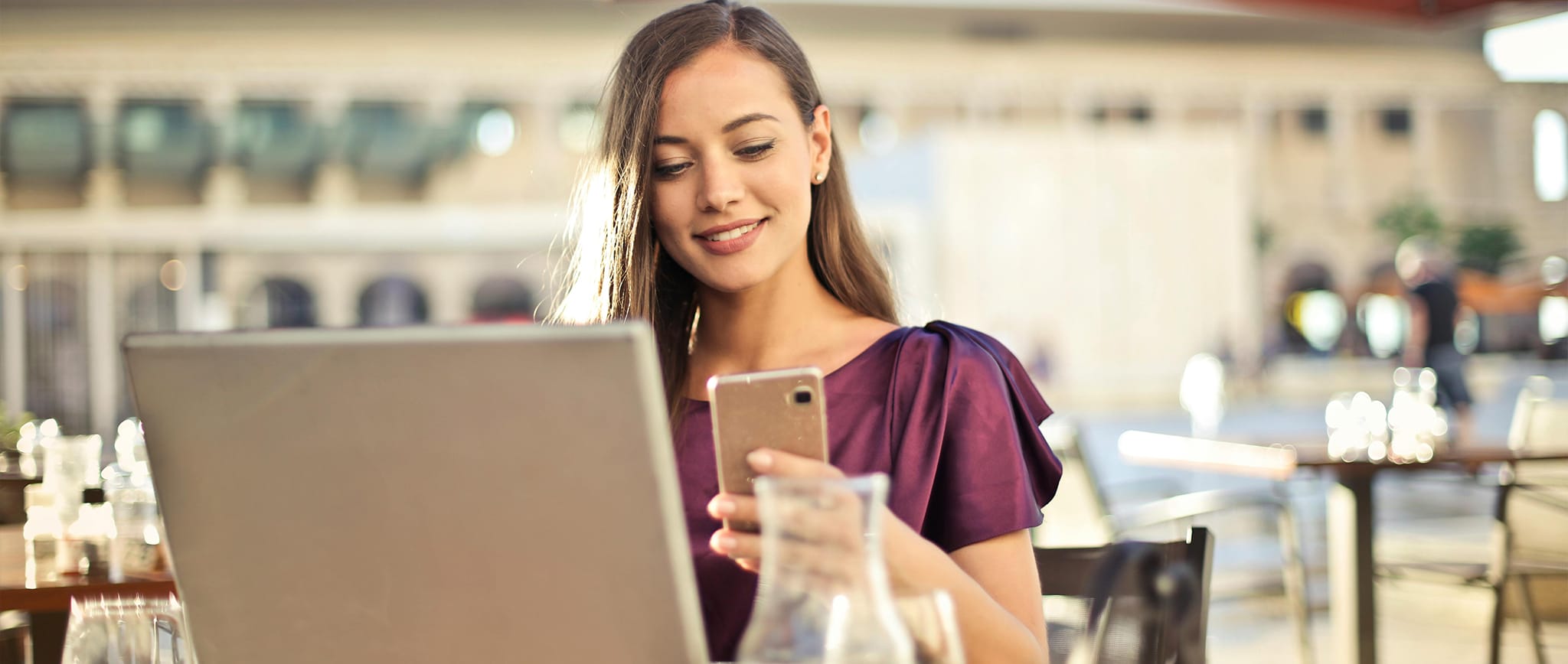Smiling woman in a terrace sitting at a dinner table, with cups and a laptop, holding a mobile phone.