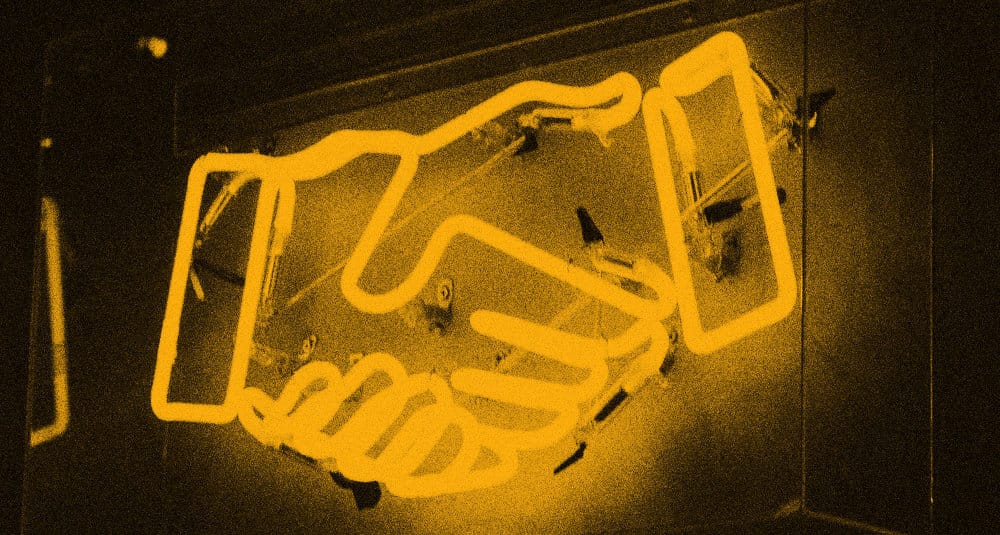 A glowing warm-orange neon sign shaped like a stylized handshake icon, mounted on a dark background.