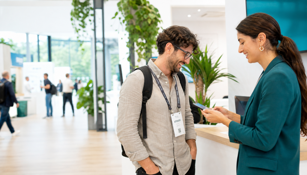 Exhibitor in a teal blazer scans a laughing attendee's badge with a smartphone at a bright booth