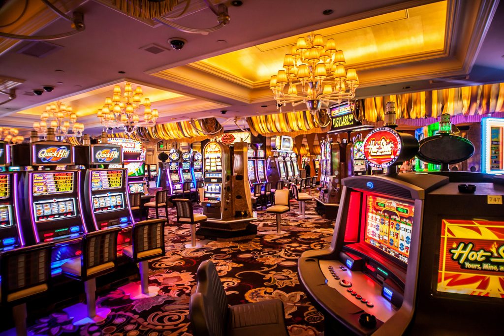 Rows of illuminated slot machines inside a lavishly decorated casino floor, featuring ornate chandeliers, patterned carpeting, and warm ambient lighting.